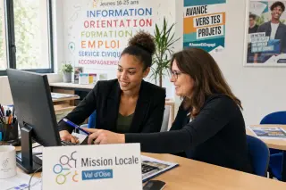 Young woman and friendly counselor working together at a desk in a bright, welcoming mission locale office, both looking at a computer screen with smiles, in an active and positive setting.
